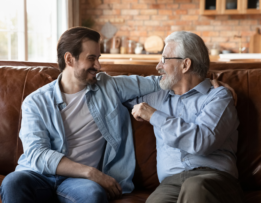 A young man and an older man sit on a brown couch looking at each other and laughing.