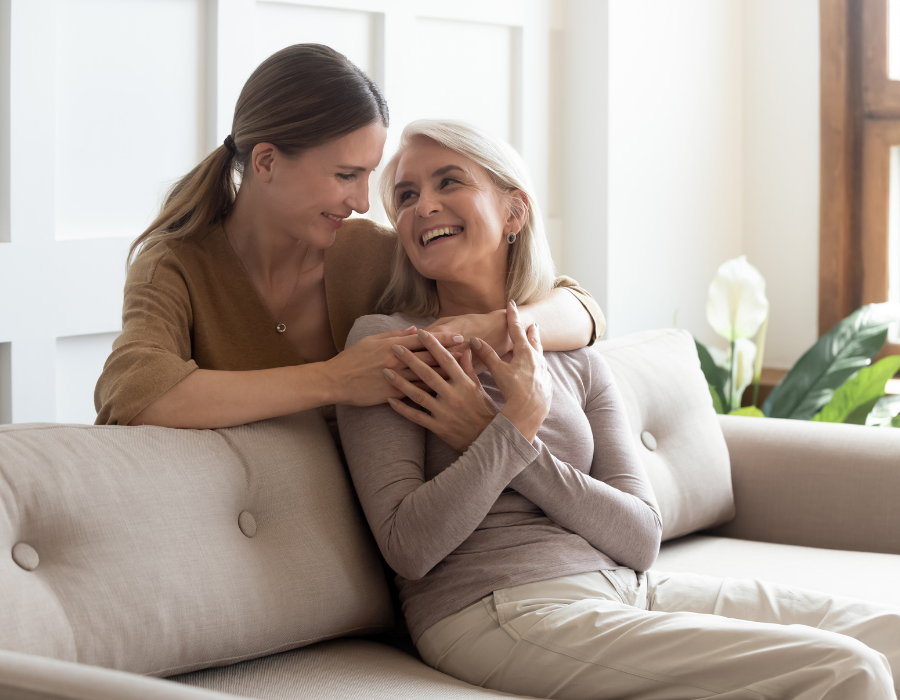 An older woman with white hair sits on a couch, her daughter hugs her from behind across the shoulders. Both are smiling