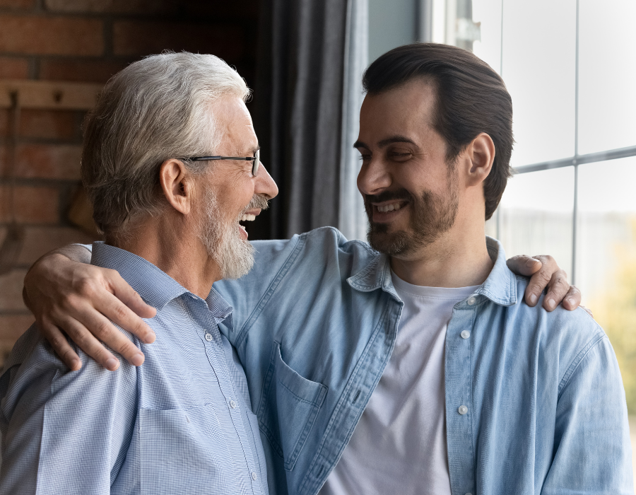 A father and son look at each other lovingly, smiling. The father is olfer with grey hair and glasses, they are in a half embrace.