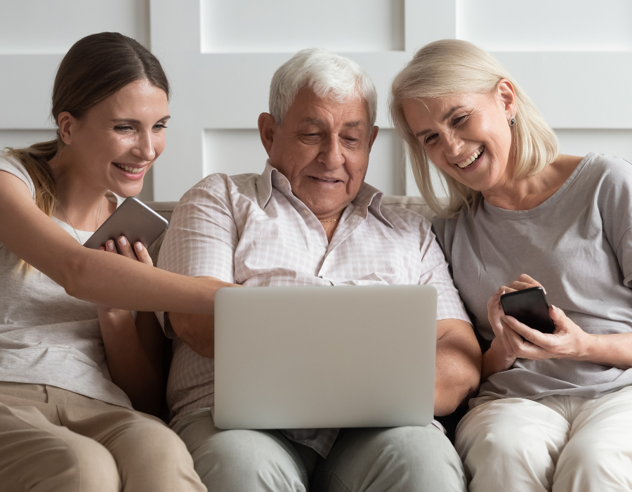 A young woman sits on the couch with her two parents. The grey haired father is in the middle holding a laptop on his lap, the mother is to his right holding a phone in her hand, They are looking at the laptop screen smiling